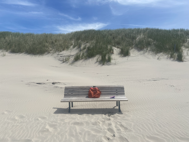 Eine Holzbank im hellen Sand auf Langeoog, davor Dünen und blauer Himmel; auf der Bank liegen eine rostrote Tasche und ein kleines Notizbuch.